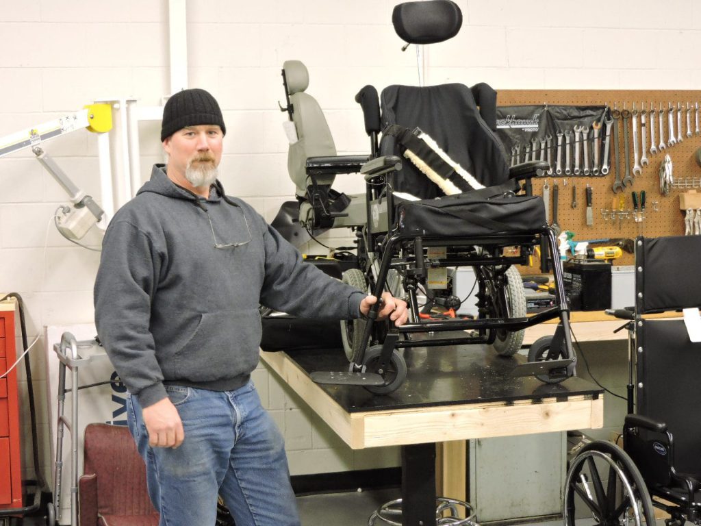Man standing at workbench with wheelchairs and tools.