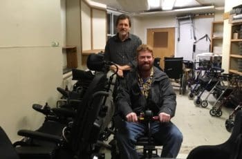 Two smiling men in a large room with mobility equipment, one is seated on a scooter.