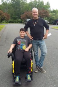Smiling boy seated in power wheelchair next to standing smiling man. 