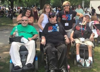 Three men seated in power wheelchairs, one using a ventolator, and three women standing behind them. 