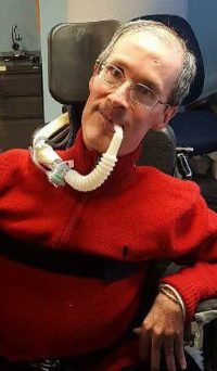 A smiling man wearing glasses seated in a power wheelchair with a head support and ventolator.
