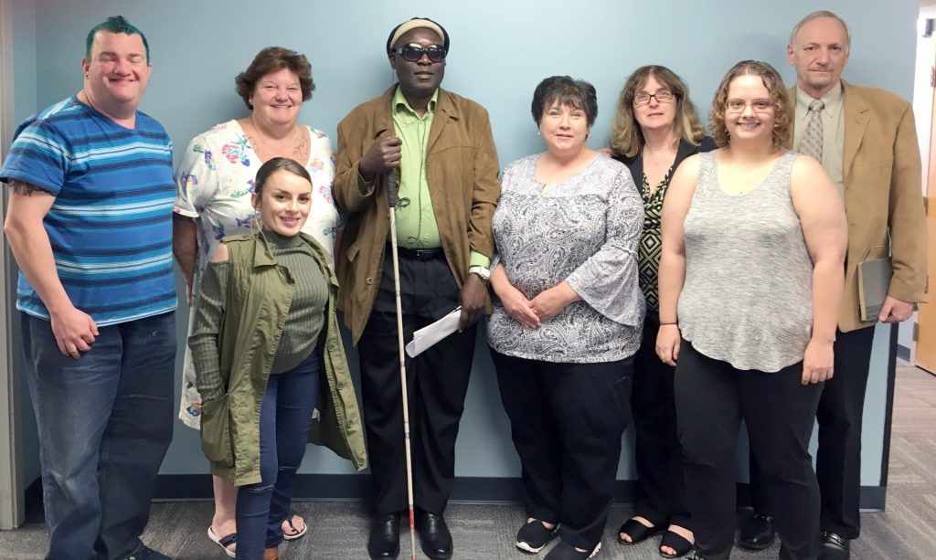 A group of standing smiling adults with and without disabilities.