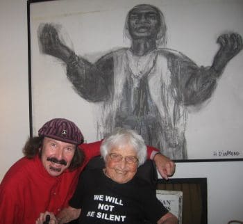 An elderly woman seated in a power chair in front of a large portrait of Sojourner Truth. A man is next to her with he arm around her. She wears a t shirt that reads We will not be silent.