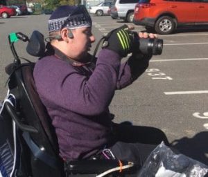 A young man holding a camera and seated in a power chair. 
