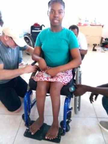 A teenage girl seated in a wheelchair while a technician kneels beside her working on her equipment.
