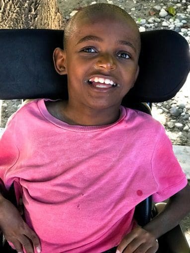 A Haitian boy seated in a wheelchair with head support smiling.