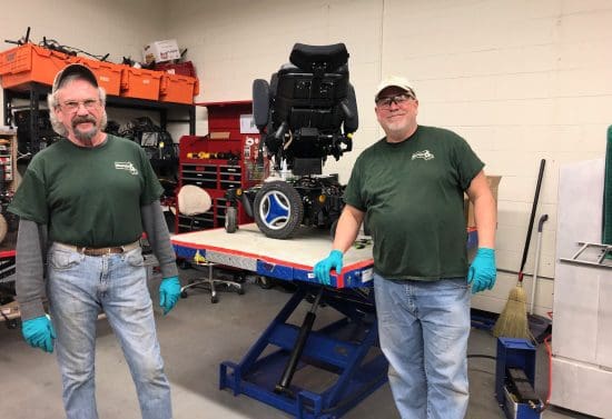 Two men smiling in front of a workbench with a power wheelchair wearing REquipment t-shirts.
