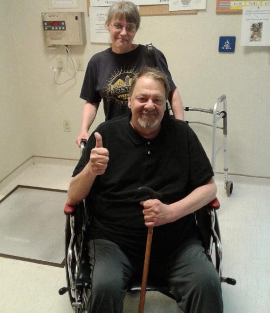 A man seated in wheelchair gives a thumbs up. His wife stands behind him smiling. They are in a hospital hallway. He holds a cane.