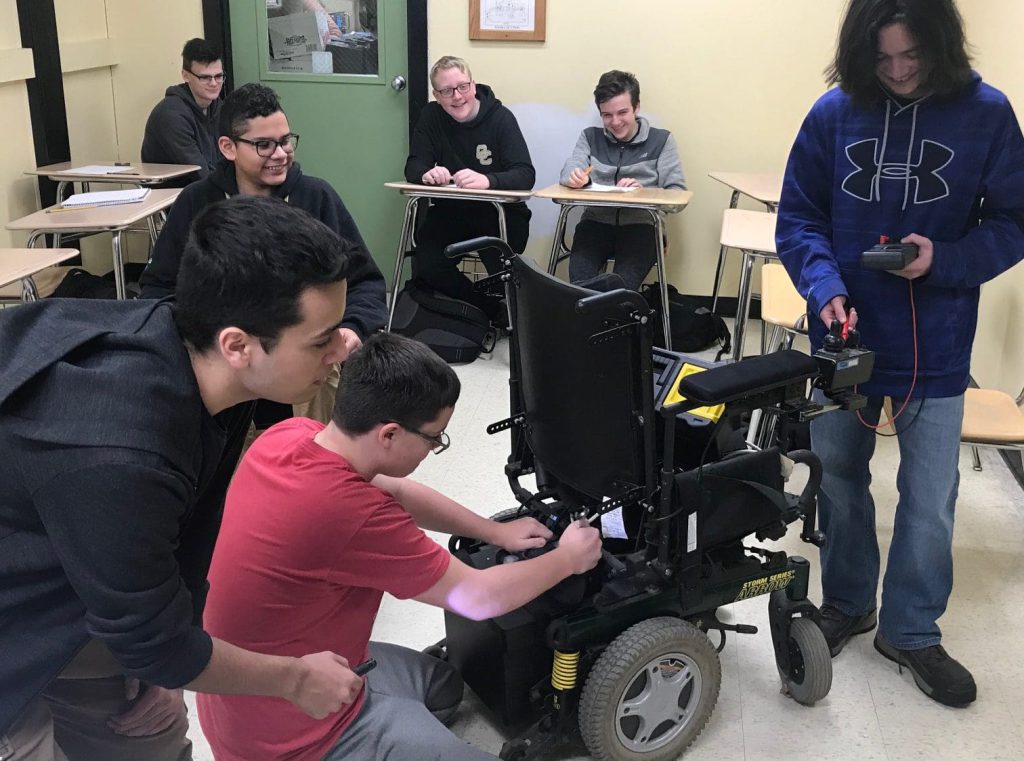 Smiling high school-aged boys repair a power wheelchair.