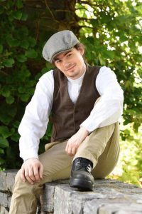 A young man wearing a cap, smiles and poses informally, seated on a stone wall.