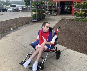 A man wearing sunglasses seated in an adapted stroller on a sidewalk outside a big box store selling plants.