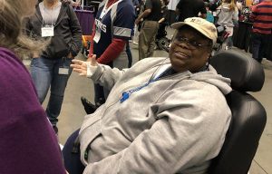 A black woman smiling seated in a power wheelchair in an exhibit hall.