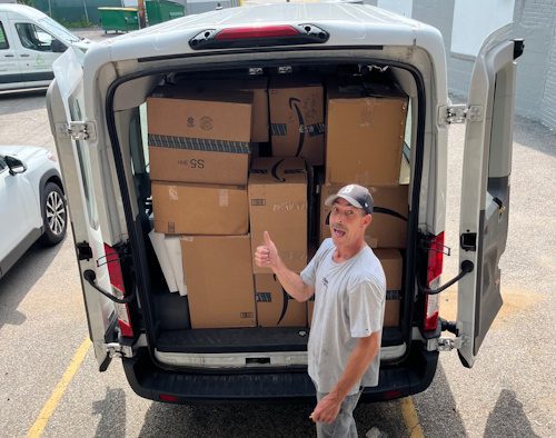 A man smiles with his mouth open (tongue protruding slightly), holding his thumb up. He is standing outdoors by the back of a van loaded with cardboard boxes.