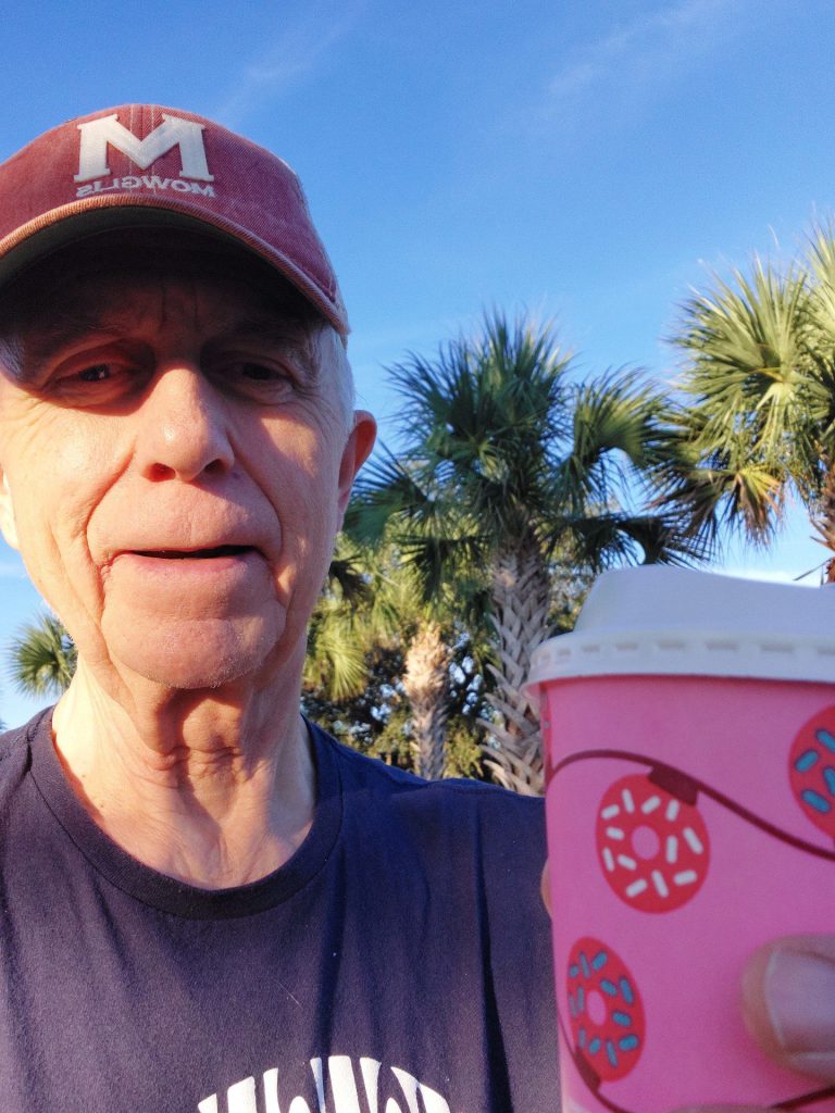 An older man smiling in the sunshine holds up a paper coffee cup with a donut design.