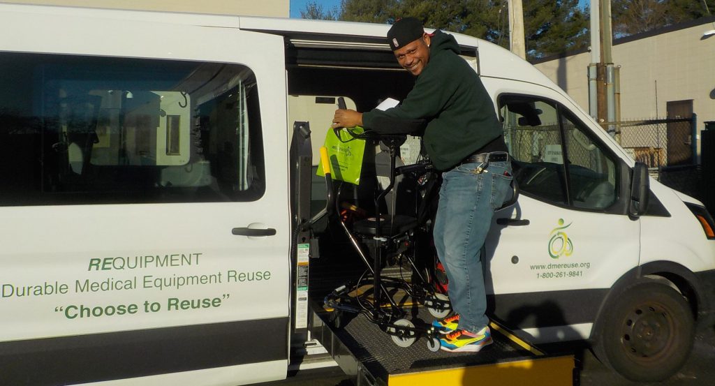 A man smiles while loading a wheelchair using a lift-equipped van that reads: REquipment Durable Medical Equipment Reuse Program. Choose To Reuse.