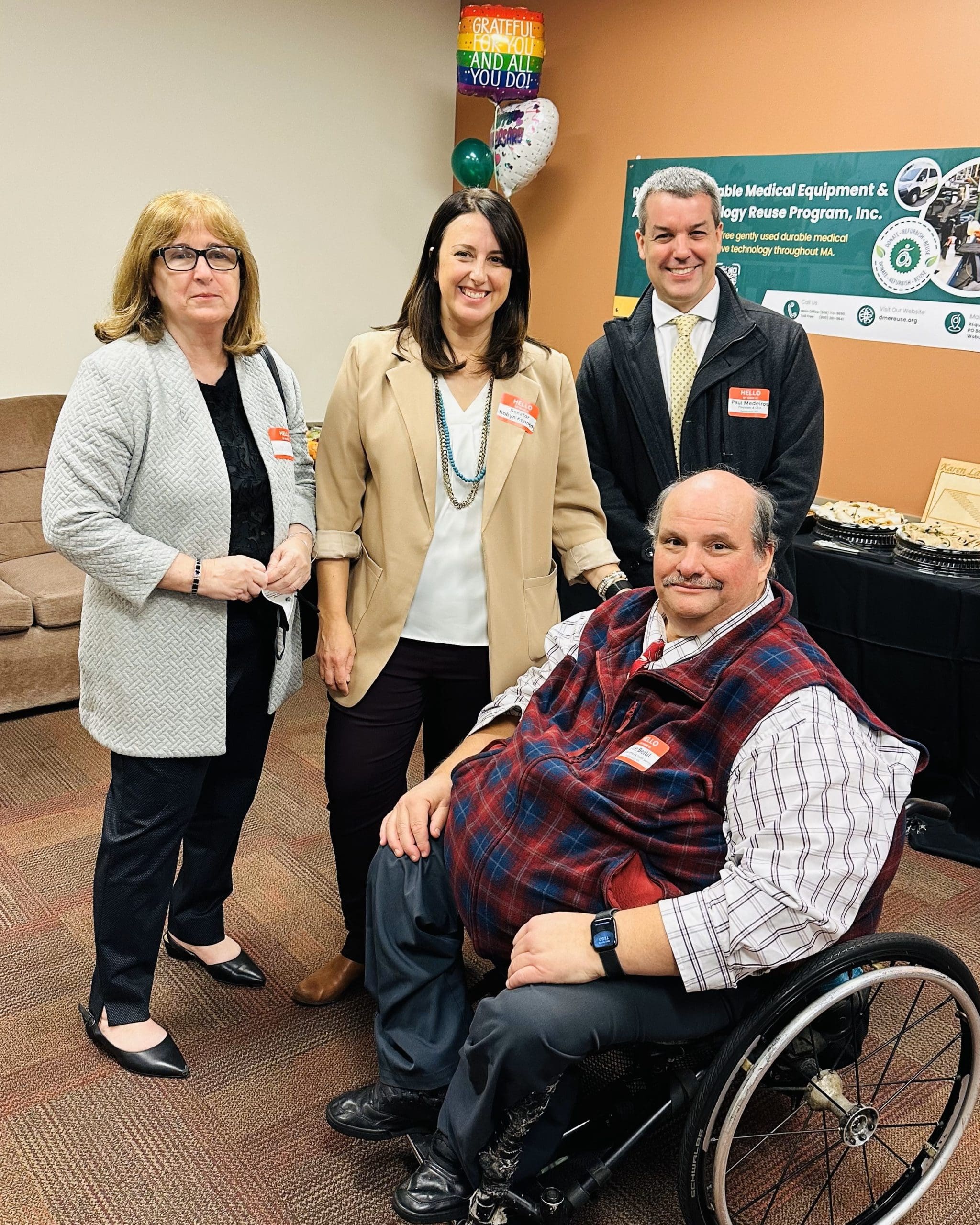 Four people smiling, three standing, a man is seated in a wheelchair.