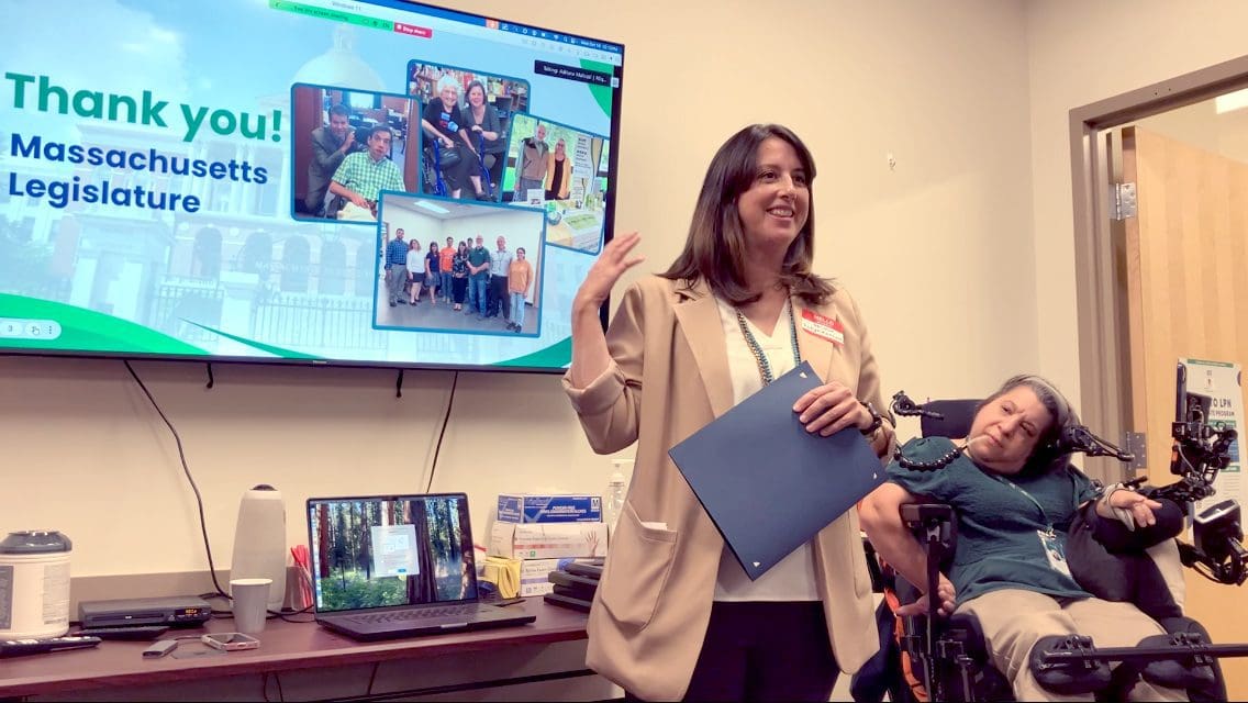 A woman smiles and gestures standing in front of a display that reads, Thank you Massachusetts Legislature.