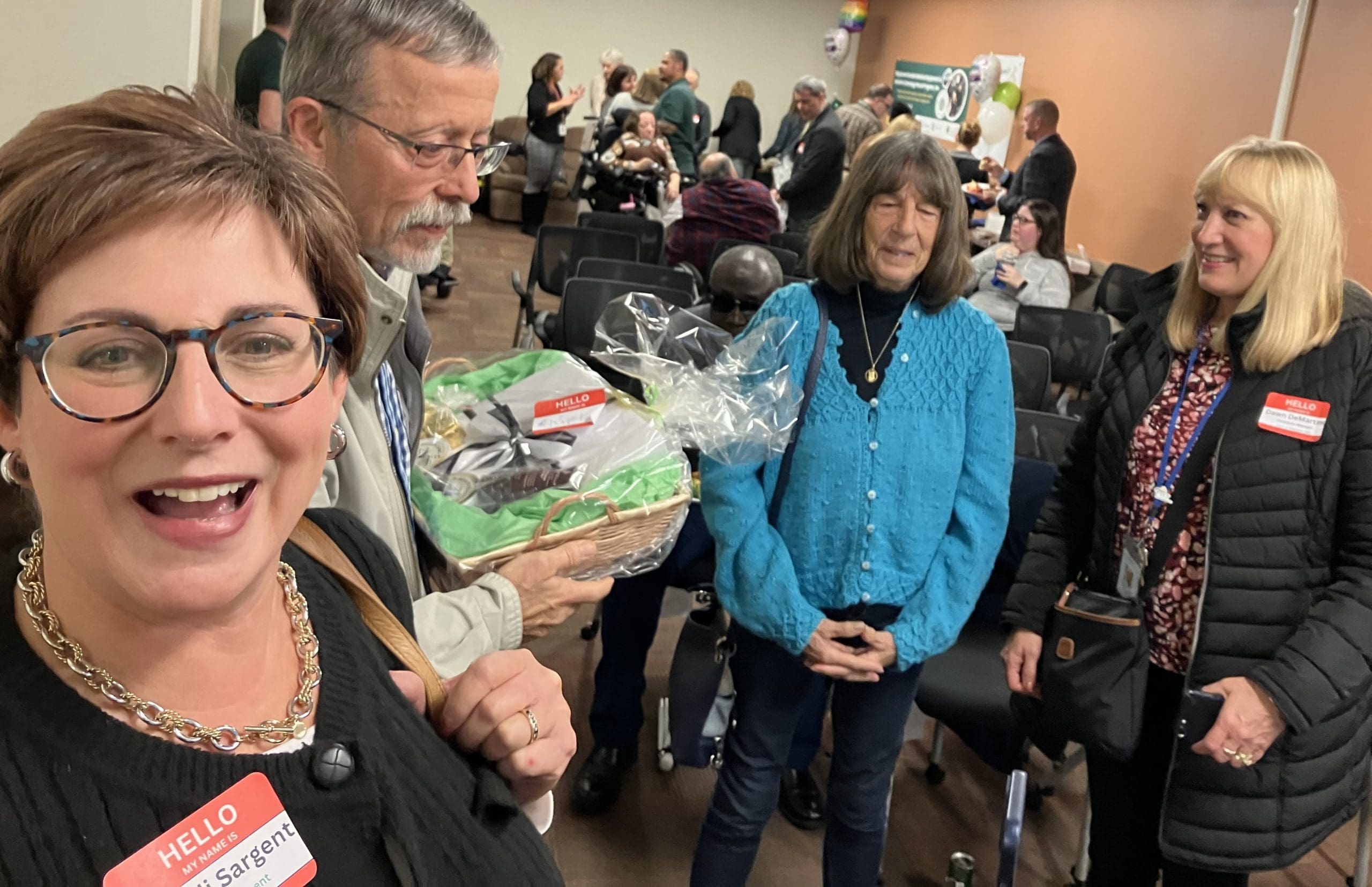 A smiling woman in the foreground of a room full of people, including a man holding a gift basket.