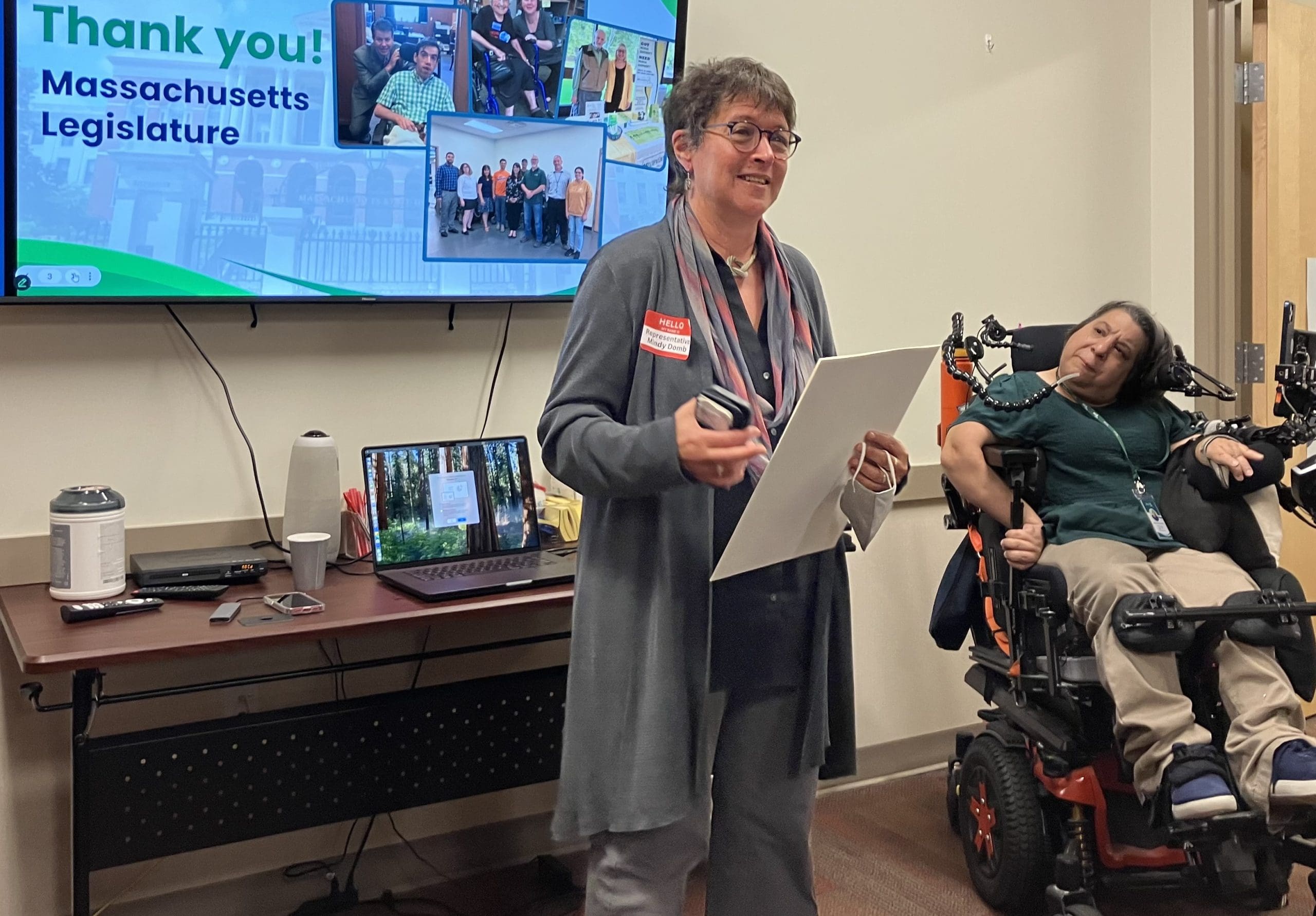 A woman stands and smiles with a large lit screen behind her with images. Another woman looks on behind seated in a power wheelchair.