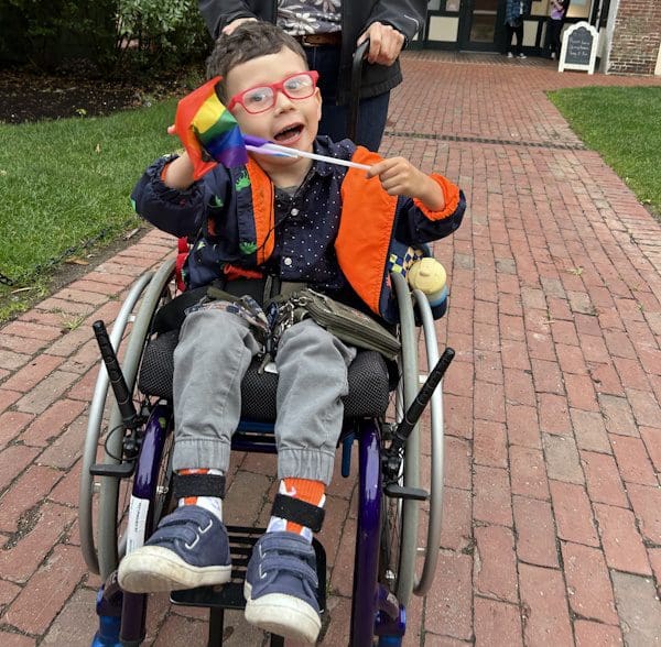A smiling young boy in a manual wheelchair with extended hand brakes holding a rainbow-colored flag.