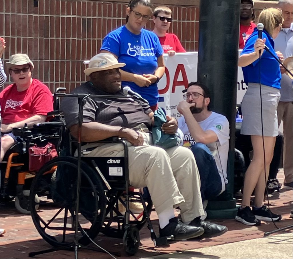 A Black man seated in a manual wheelchair speaks into a microphone surrounded by other people seated and standing.