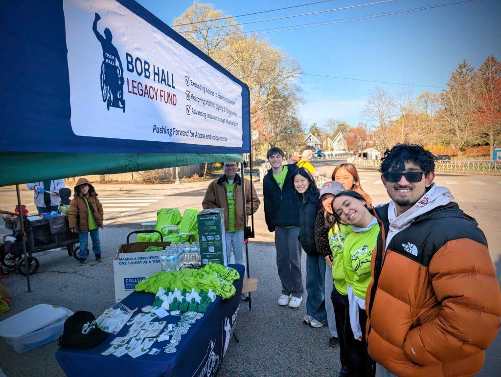 Smiling young adults with a table of race day swag and a Bob Hall Legacy Fund banner.