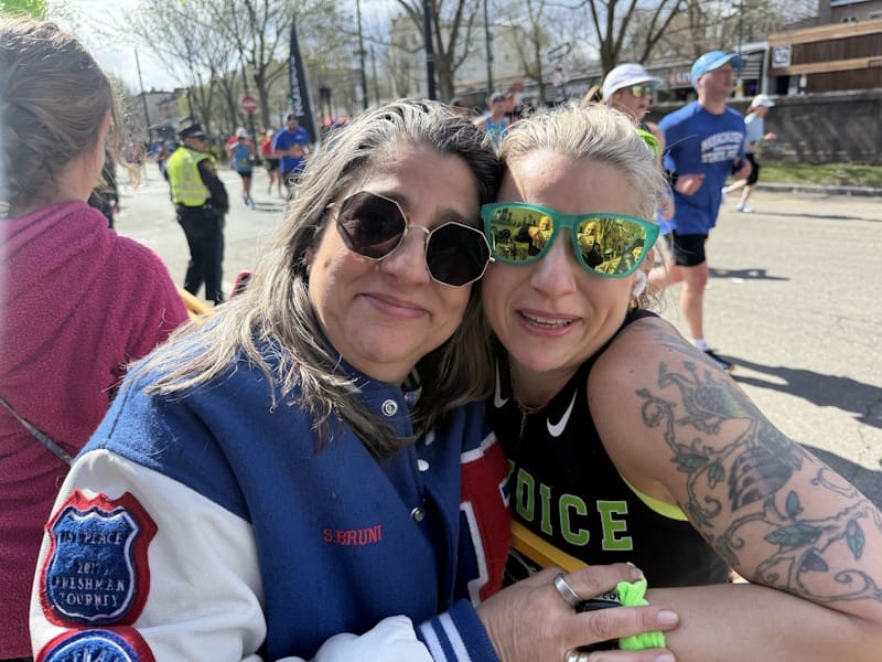 Two women cheek-to-cheek wearing sunglasses, smile for the camera outdoors backdropped by runners.
