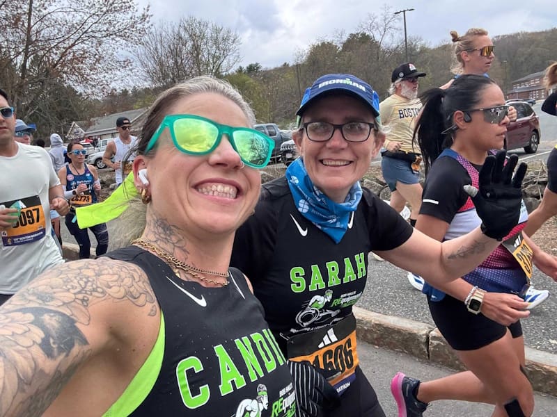 Two women beam smiles for a selfie in a crowd of runners, names on their shirts. Candice is taking it with her tattooed arm wearing mirrored green sunglasses. Sarah wears an Ironman cap and waves her gloved hand.