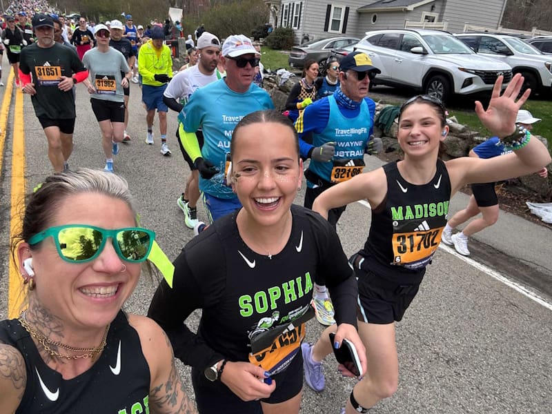 Three women smile broadly for the camera while running wearing team shirts, more runners behind them.