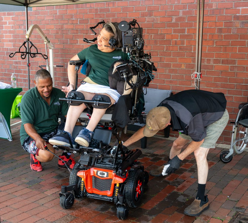 A woman laughing waiting in her power wheelchair while two men clean it.