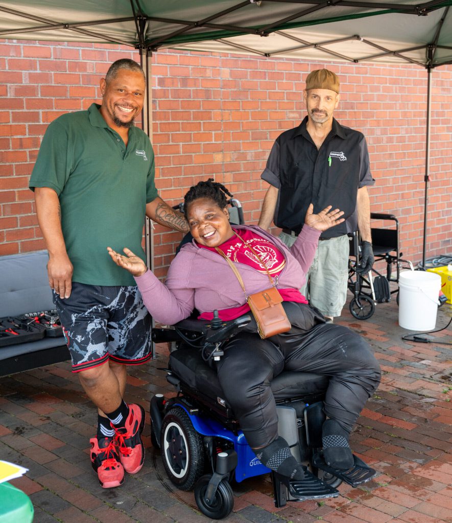 A woman beams in her wheelchair flanked by two smiling men under a tent outdoors.