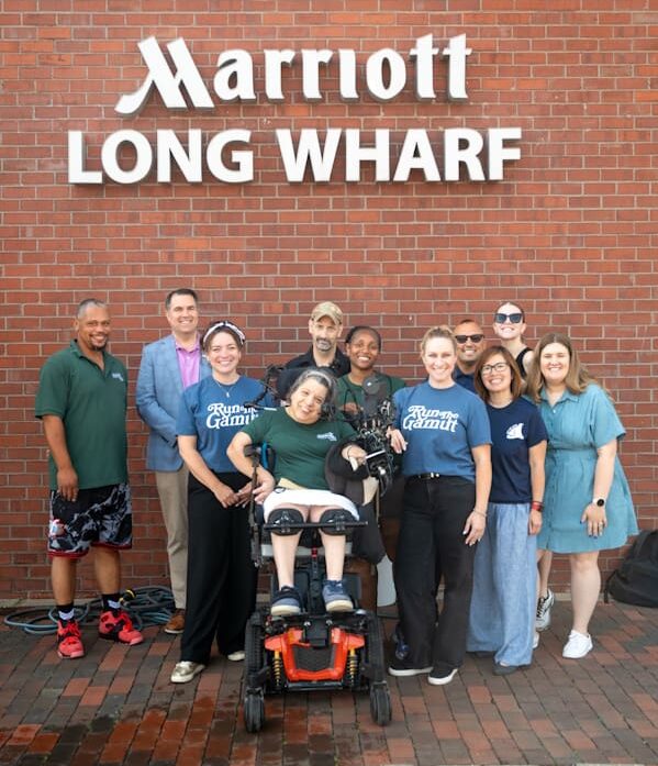 A woman in a power wheelchair surrounded by ten adults, some in REquipment and Run the Gamut shirts, pose in front of a brick wall with signage for the Marriott Long Wharf.