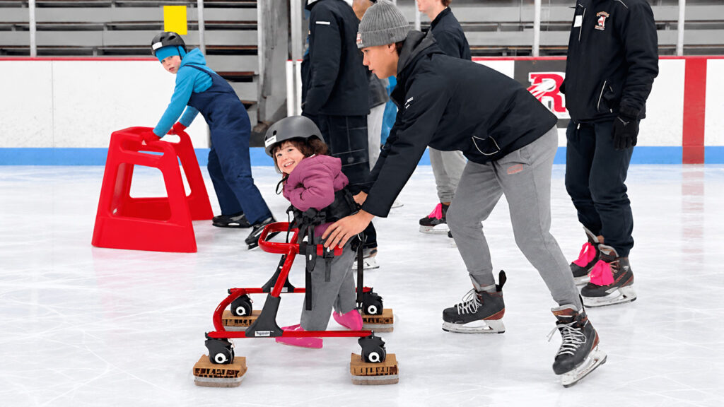 Hazel smiles while using her gait trainer adapted for ice skating as a teenage volunteer pushes her across the rink.