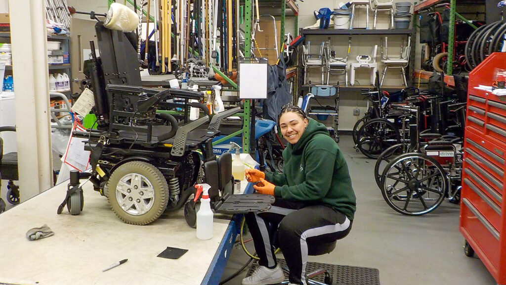 A REquipment staff member cleans a power wheelchair in a workshop surrounded by wheelchairs and mobility equipment.