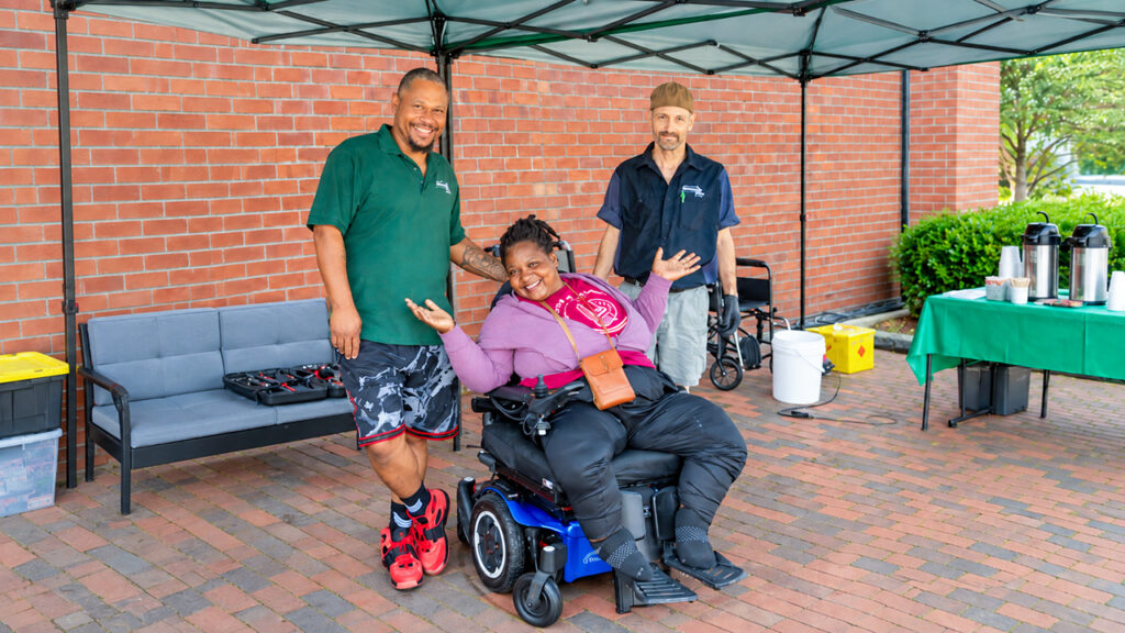 A community member smiles in her power wheelchair with REquipment technicians Anthony Calderon-Diaz and Jim Riel at a Wheelchair Wash in Boston.