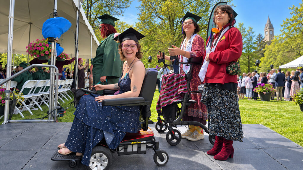 Rue Fishman smiles in a power wheelchair with fellow graduates at Hampshire College commencement.